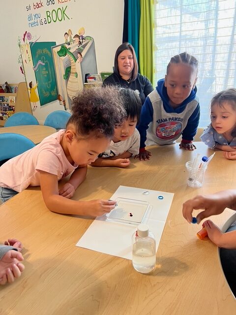 Children sitting around a table, focused on a child who is drawing, showcasing creativity and collaboration.