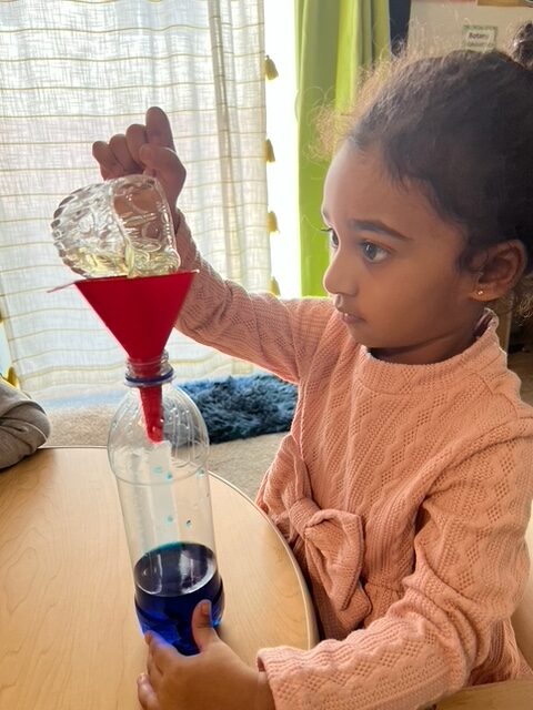 A young girl is engaged in a hands-on activity, pouring liquid into a funnel with focused concentration.